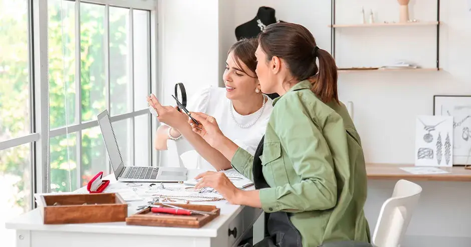 2 women discussing, 1 is holding up a jewelry sample, they are in an office studio setting with jewelry item throughout the room