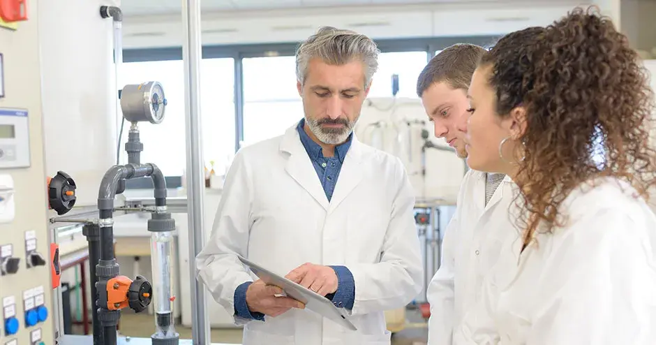 Bearded man in lab coat showing clipboard to two colleagues (both in lab coats, a man and woman) in a industrial setting